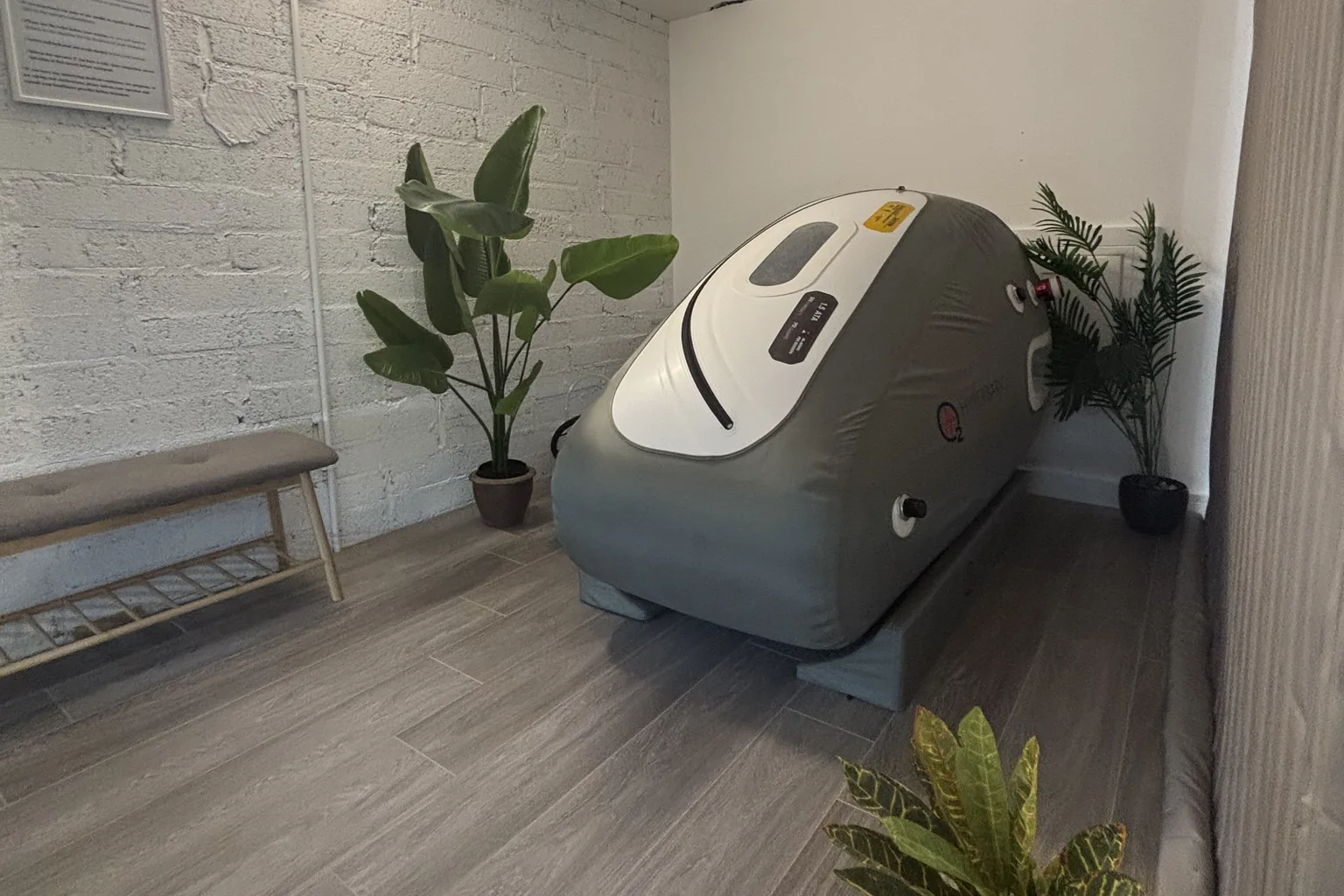 The chamber room: a grey soft-shell hyperbaric chamber on a wood-plank floor, a bench and tall plants along the white brick walls