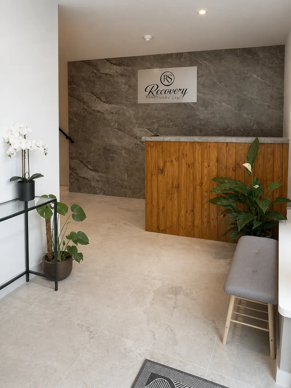 Reception: a honey-pine desk against a grey stone feature wall carrying the Recovery Sanctuary sign, orchids and a monstera on pale stone tile
